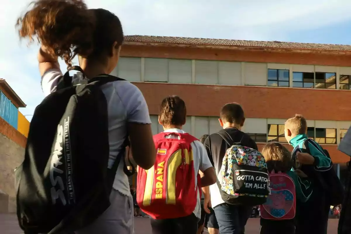 Un grupo de niños con mochilas camina hacia un edificio escolar de ladrillo bajo un cielo despejado. Un grupo de niños con mochilas camina hacia un edificio escolar de ladrillo bajo un cielo despejado.
