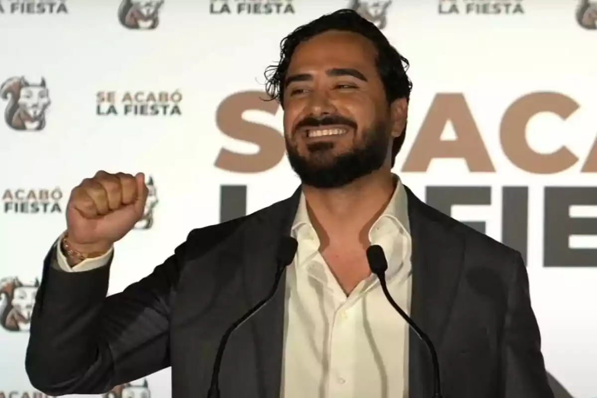 Plano medio de Alvise Pérez sonriendo y alzando un puño con un fondo lleno de logos de su partido Se Acabó La Fiesta