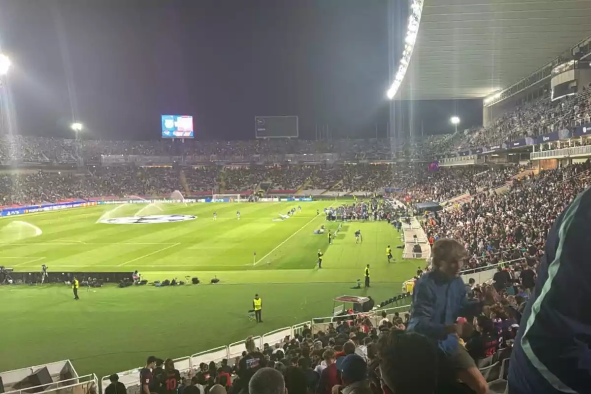 Un estadio de fútbol lleno de espectadores durante un partido nocturno con el campo iluminado y aspersores en funcionamiento. Un estadio de fútbol lleno de espectadores durante un partido nocturno con el campo iluminado y aspersores en funcionamiento.