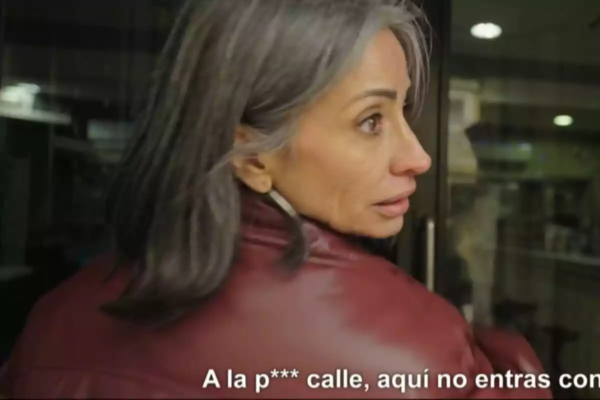 Una mujer de cabello canoso y chaqueta roja está mirando hacia atrás con una expresión seria, mientras se encuentra frente a una puerta de vidrio en Apatrullando.