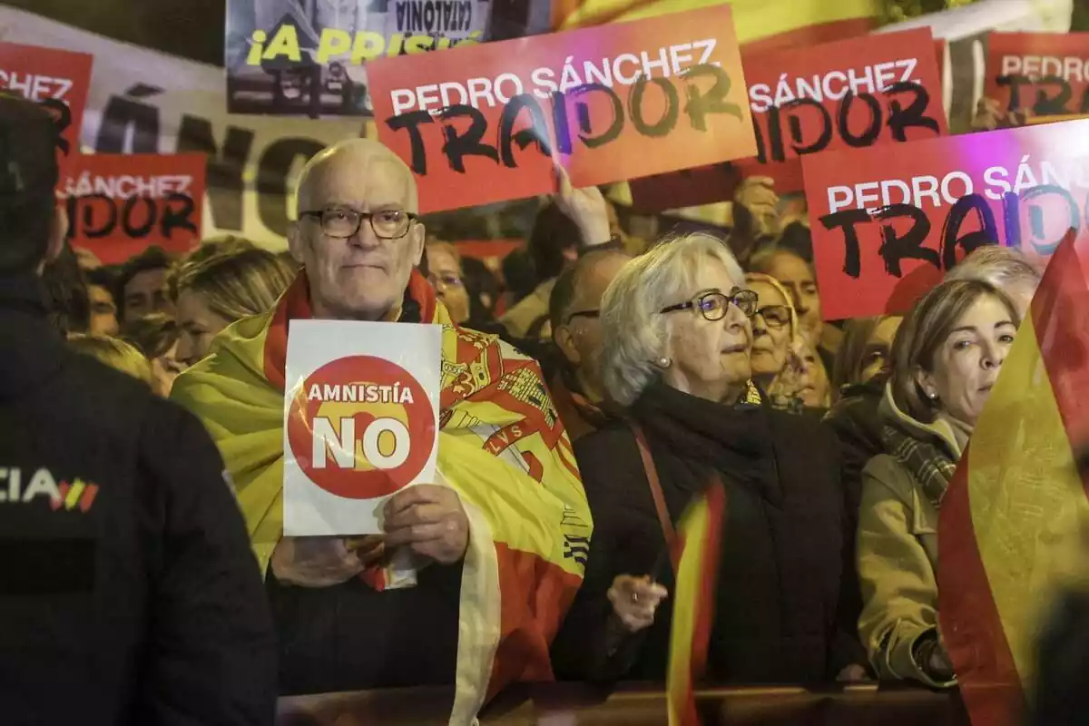 Un señor con una bandera de España en modo capa sujeta un cartel con la frase 'amnistía no'. Detrás de él, varias personas muestras carteles con el lema 'Pedro Sánchez traidor'