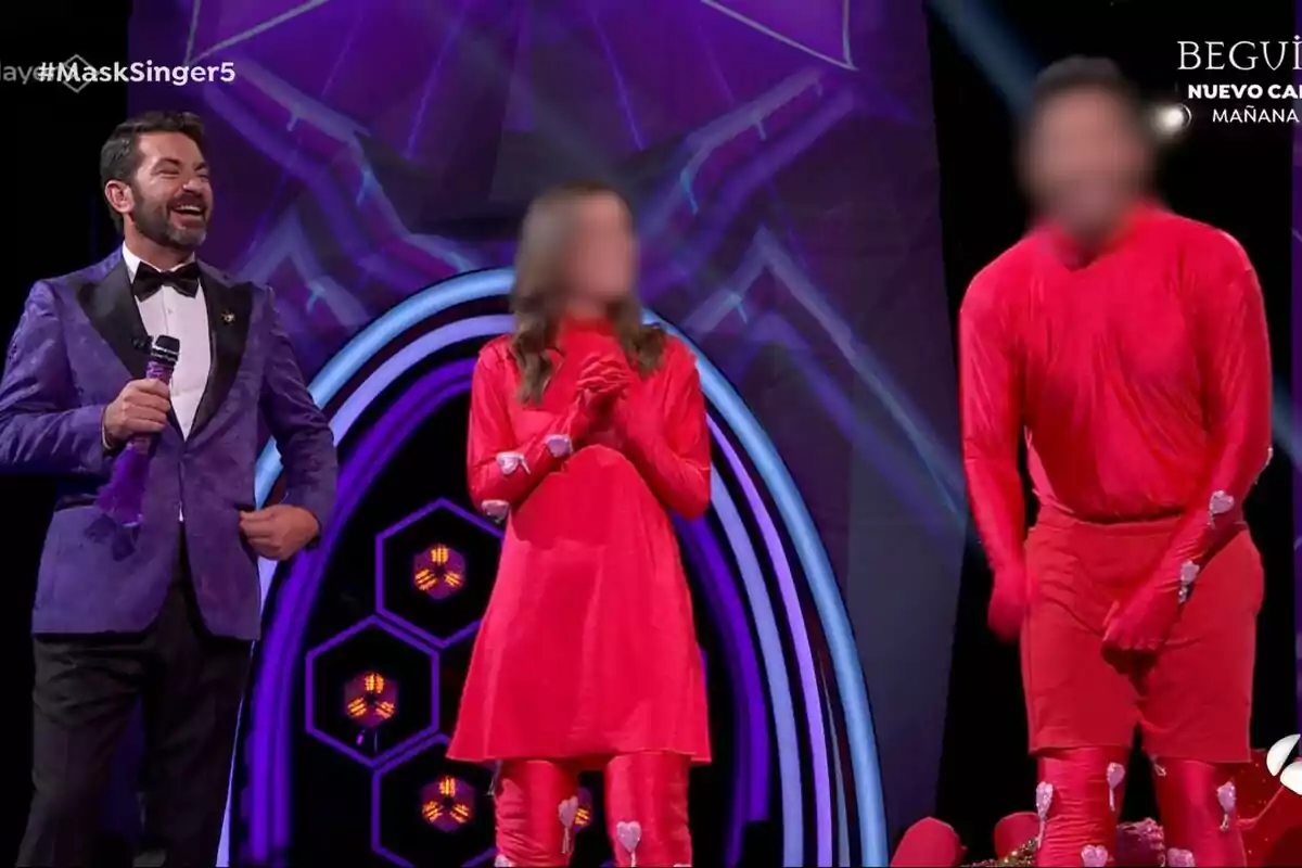 Arturo Valls sonriente con un traje morado sostiene un micrófono mientras dos personas disfrazadas de rojo están a su lado en un escenario iluminado en Mask Singer.