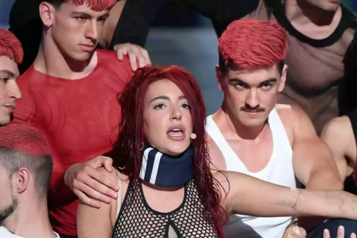 Grupo de personas con el cabello teñido de rojo, Ana Guerra al centro lleva collarín y micrófono, rodeada por hombres con ropa ajustada en Tu Cara Me Suena Grupo de personas con el cabello teñido de rojo, Ana Guerra al centro lleva collarín y micrófono, rodeada por hombres con ropa ajustada en Tu Cara Me Suena