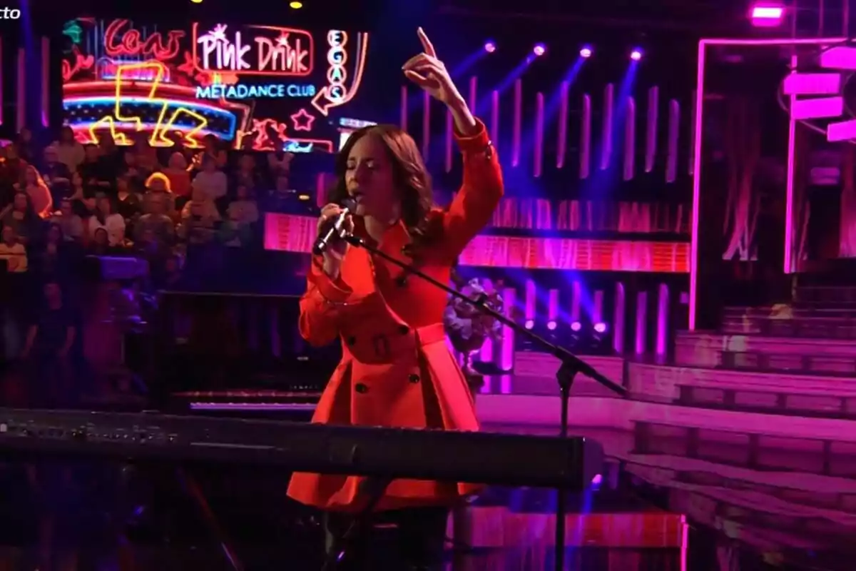 Ana Guerra con un abrigo rojo canta en un escenario iluminado con luces de neón y un público al fondo en Tu Cara Me Suena. Ana Guerra con un abrigo rojo canta en un escenario iluminado con luces de neón y un público al fondo en Tu Cara Me Suena.