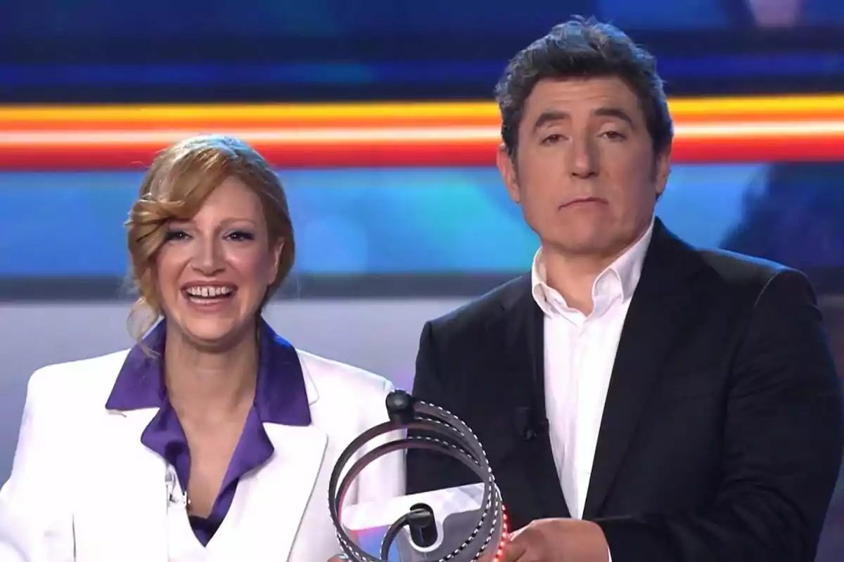 Ana Guerra y Manel Fuentes sonríen mientras sostienen el pulsador de Tu Cara Me Suena en un escenario iluminado.