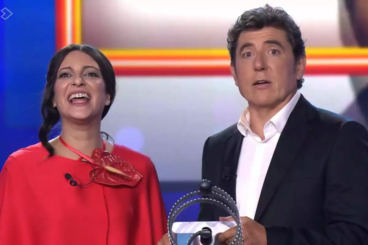 Dos presentadores en Tu Cara Me Suena, Ana Guerra sonríe ampliamente y Manel Fuentes luce sorprendido, ambos están de pie frente a una máquina o dispositivo circular