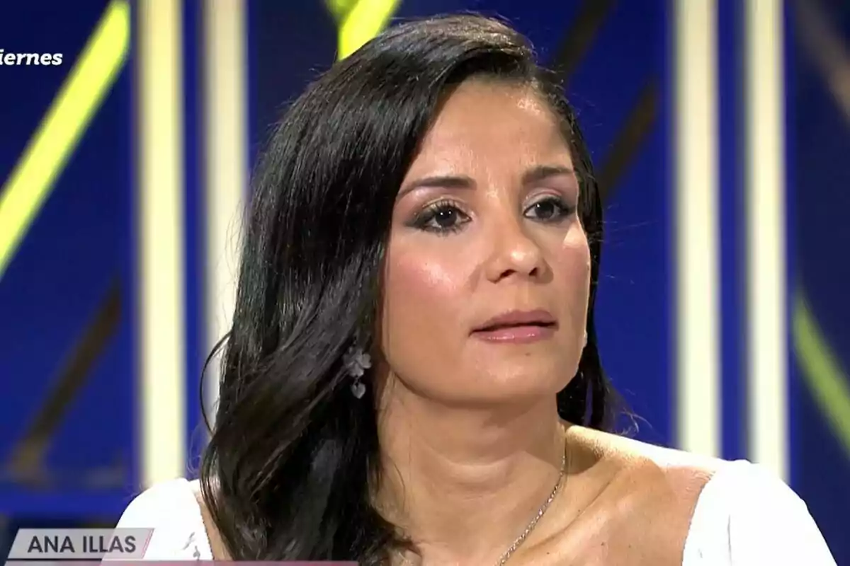 Captura de Ana Herminia con cabello oscuro y largo, usando un vestido blanco, aparece en el plató de De Viernes con un fondo azul y amarillo.