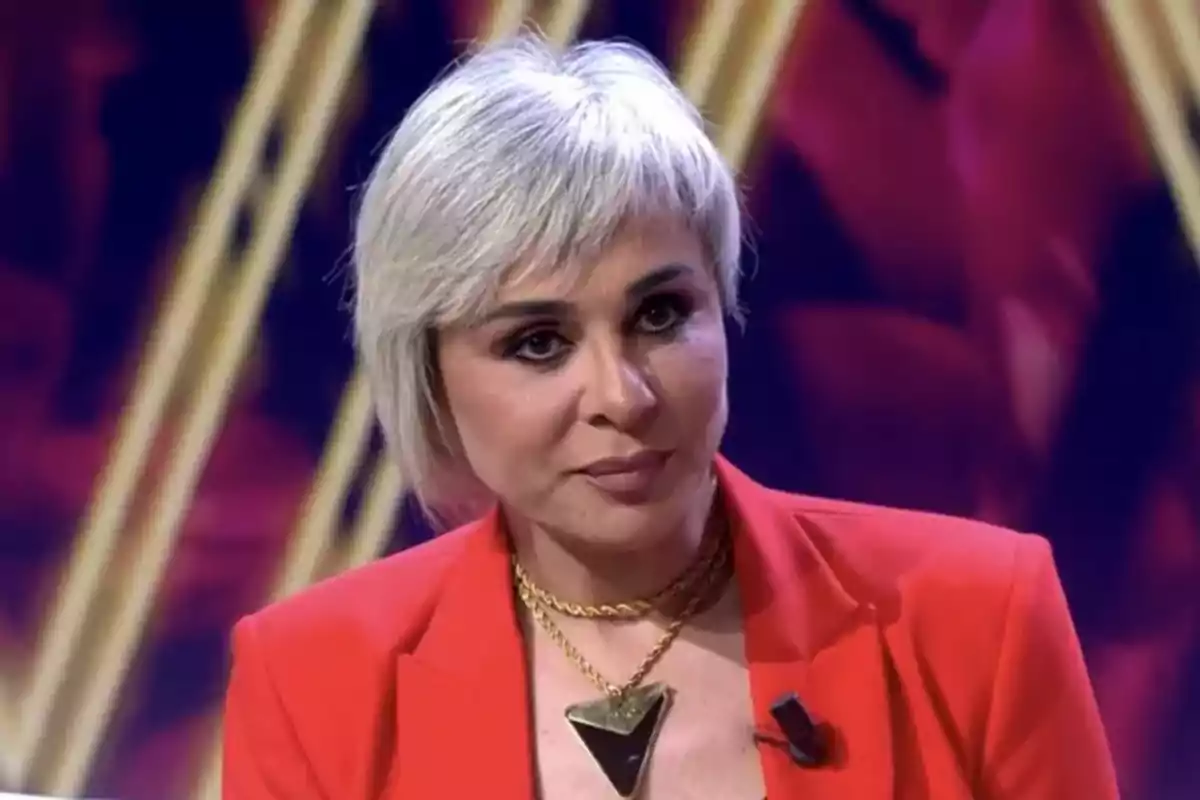 Ana María Aldón con cabello corto y rubio vestida con un traje rojo y un collar dorado en un ambiente iluminado. Ana María Aldón con cabello corto y rubio vestida con un traje rojo y un collar dorado en un ambiente iluminado.