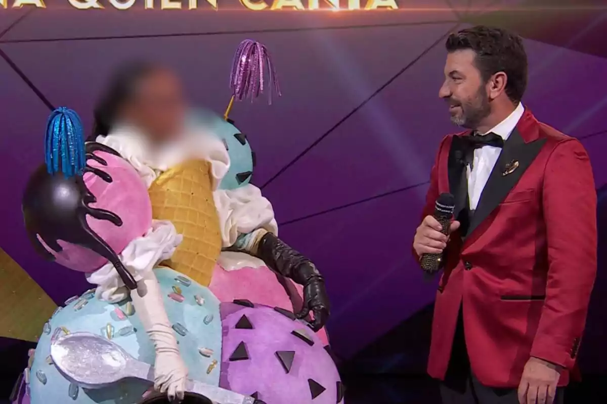 Ana Peleteiro disfrazada de helado con conos y bolas de colores está junto a un presentador que lleva un traje rojo y sostiene un micrófono en Mask Singer.