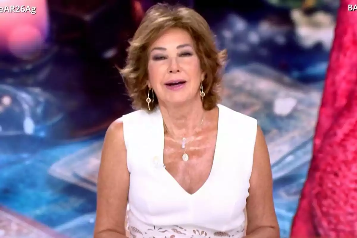 Ana Rosa Quintana con cabello corto y castaño, vestida con una blusa blanca sin mangas, está sonriendo frente a una cámara en un entorno de estudio con un fondo desenfocado en TardeAR.