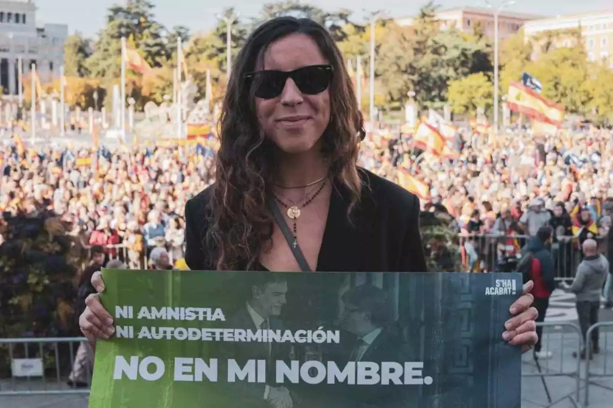 Andrea Llopart sonriendo con gafas de sol y sujetando un cartel verdo con el lema 'ni amnistía ni autodeterminación, no en mi nombre'. De fondo, una concentración con centenares de personas con banderas españolas