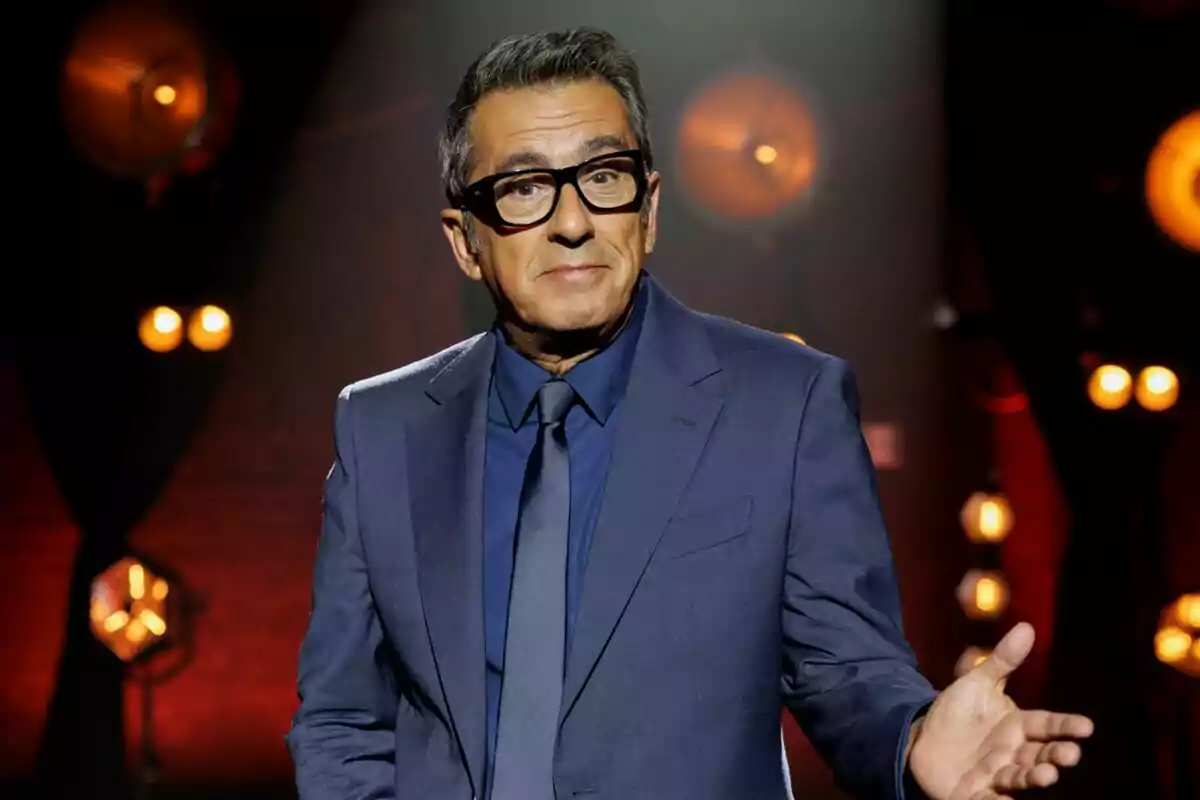 Andreu Buenafuente con gafas y traje azul oscuro gesticula con una mano en un escenario iluminado con luces cálidas en Futuro Imperfecto.