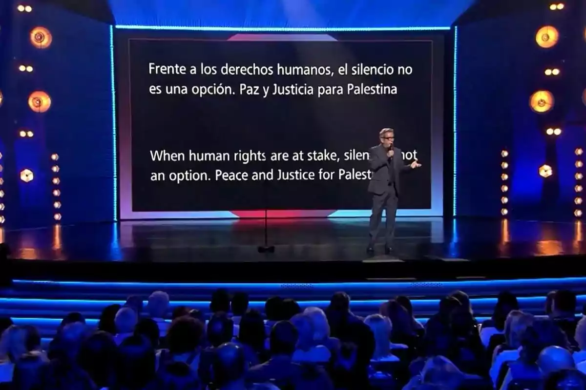 Andreu Buenafuente en un escenario de Futuro Imperfecto con una pantalla grande detrás que muestra un mensaje sobre derechos humanos y justicia para Palestina, mientras el público observa. Andreu Buenafuente en un escenario de Futuro Imperfecto con una pantalla grande detrás que muestra un mensaje sobre derechos humanos y justicia para Palestina, mientras el público observa.