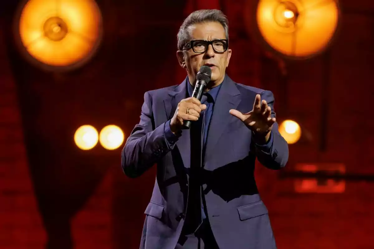 Andreu Buenafuente con gafas y traje azul sostiene un micrófono en un escenario iluminado con luces cálidas en Futuro Imperfecto. Andreu Buenafuente con gafas y traje azul sostiene un micrófono en un escenario iluminado con luces cálidas en Futuro Imperfecto.