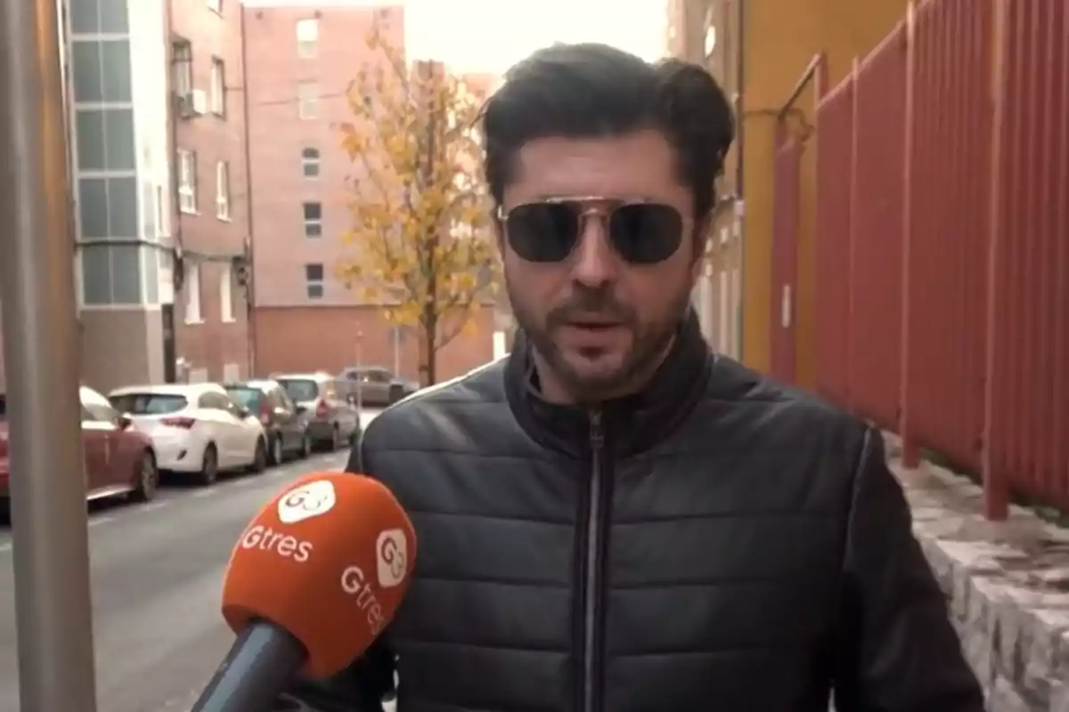 Ángel Cristo, con chaqueta negra y gafas de sol, es entrevistado en la calle por un micrófono naranja de Gtres