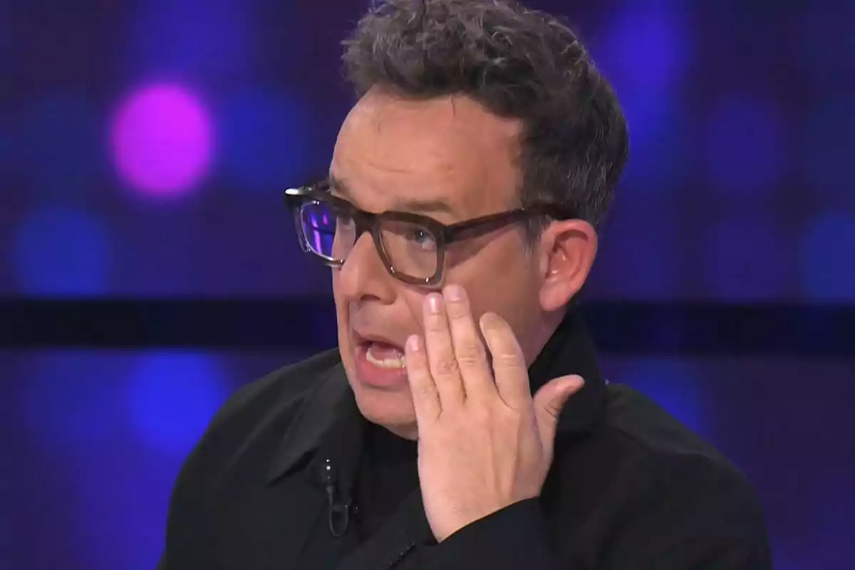 Àngel Llàcer con gafas y expresión emocional se toca la cara mientras habla en un entorno iluminado con luces de fondo en Tu Cara Me Suena.