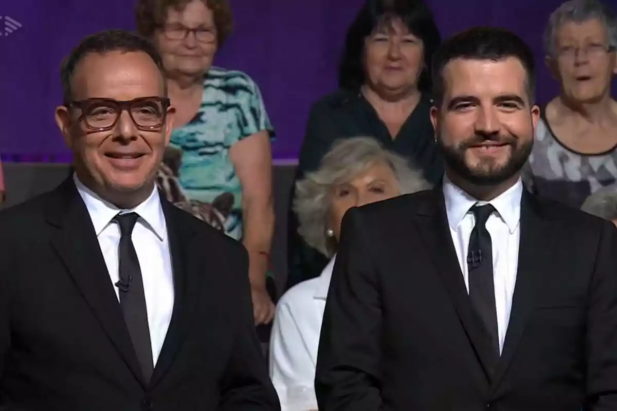 Àngel Llàcer y Ricard Ustrell vestidos de traje y corbata negra sonríen frente a una audiencia en un estudio de televisión de Col·lapse. Àngel Llàcer y Ricard Ustrell vestidos de traje y corbata negra sonríen frente a una audiencia en un estudio de televisión de Col·lapse.