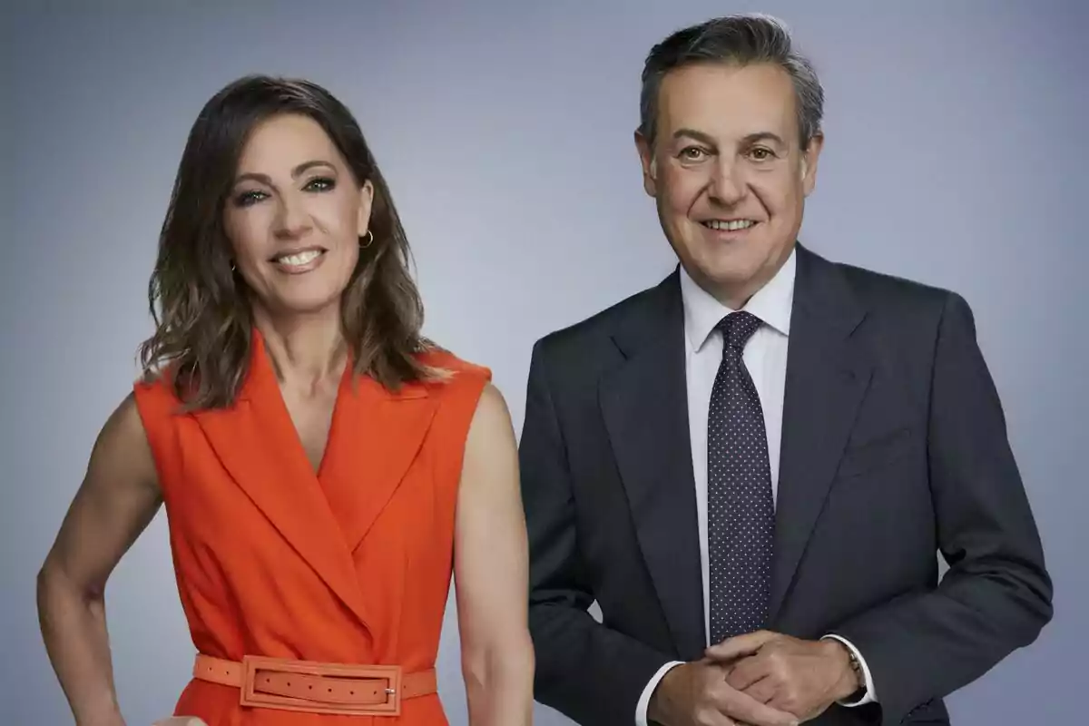 Ángeles Blanco y Pepe Ribagorda posan sonrientes frente a un fondo gris, una mujer con vestido naranja sin mangas y un hombre con traje oscuro y corbata. Ángeles Blanco y Pepe Ribagorda posan sonrientes frente a un fondo gris, una mujer con vestido naranja sin mangas y un hombre con traje oscuro y corbata.