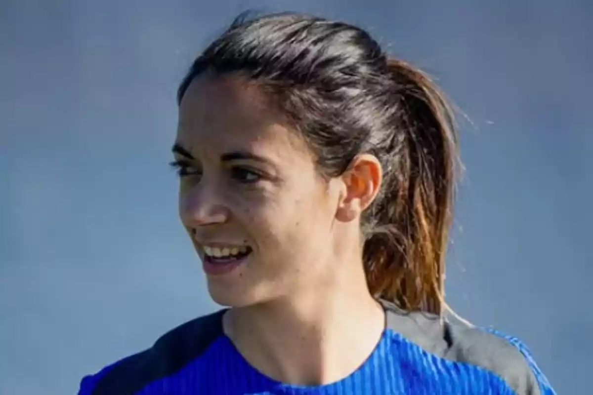 Una persona con cabello recogido y camiseta azul sonríe mientras mira hacia un lado. Una persona con cabello recogido y camiseta azul sonríe mientras mira hacia un lado.
