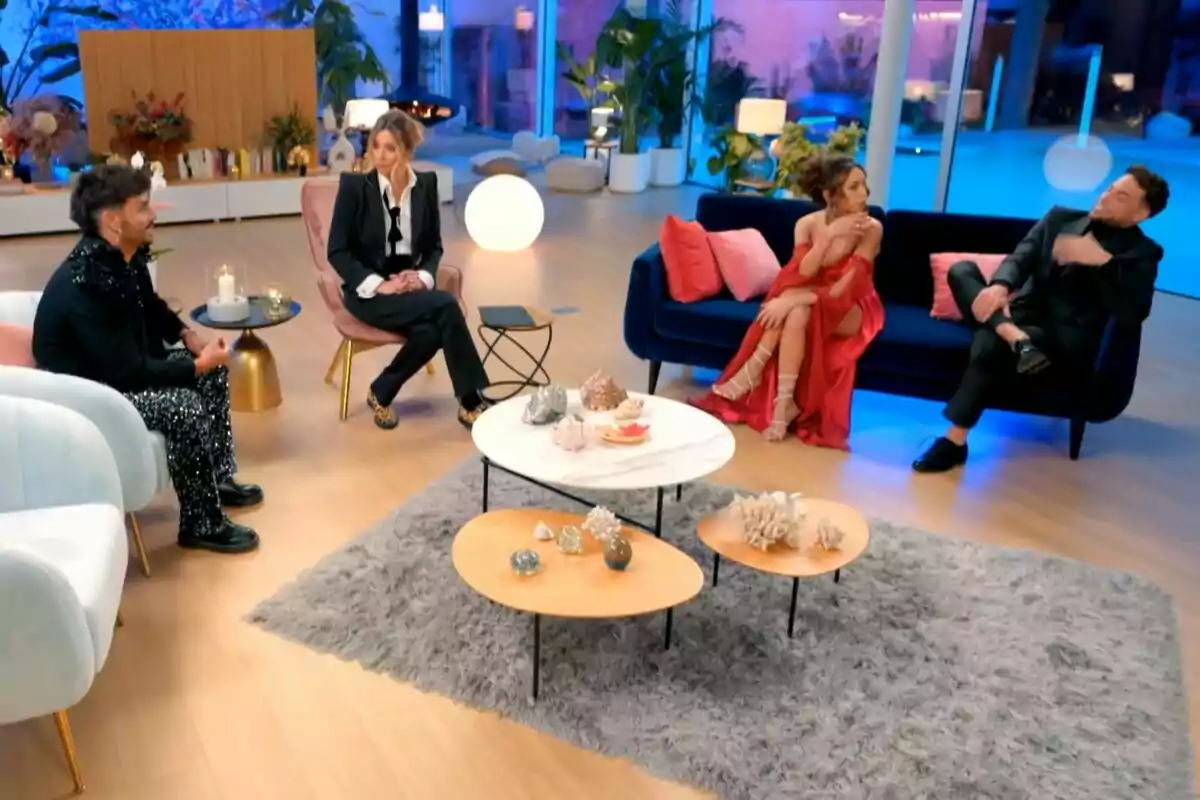 Un grupo de personas formado por Manuel González, Sandra Barneda, Anita Williams y Montoya sentadas en un salón moderno con decoración elegante y luces azules en La Isla de las Tentaciones. Un grupo de personas formado por Manuel González, Sandra Barneda, Anita Williams y Montoya sentadas en un salón moderno con decoración elegante y luces azules en La Isla de las Tentaciones.