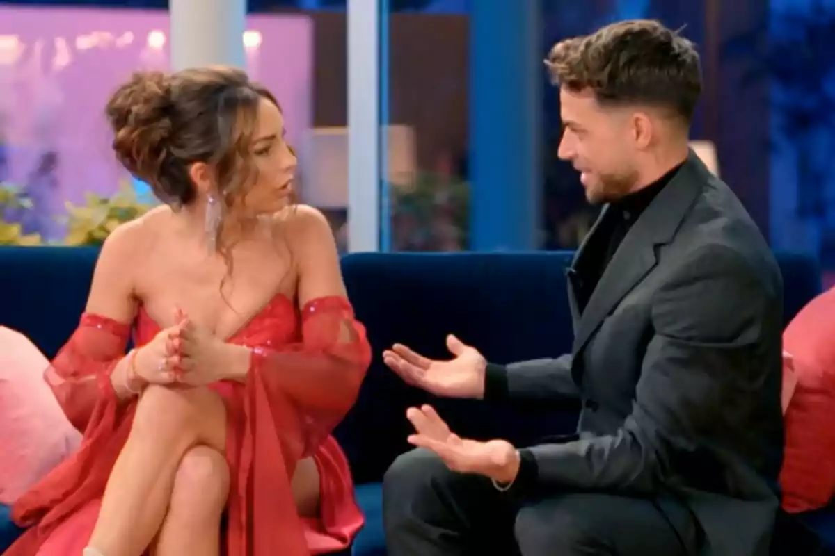 Anita Williams y Montoya sentadas en un sofá conversando intensamente, una mujer con vestido rojo y un hombre con traje oscuro en La Isla de las Tentaciones.