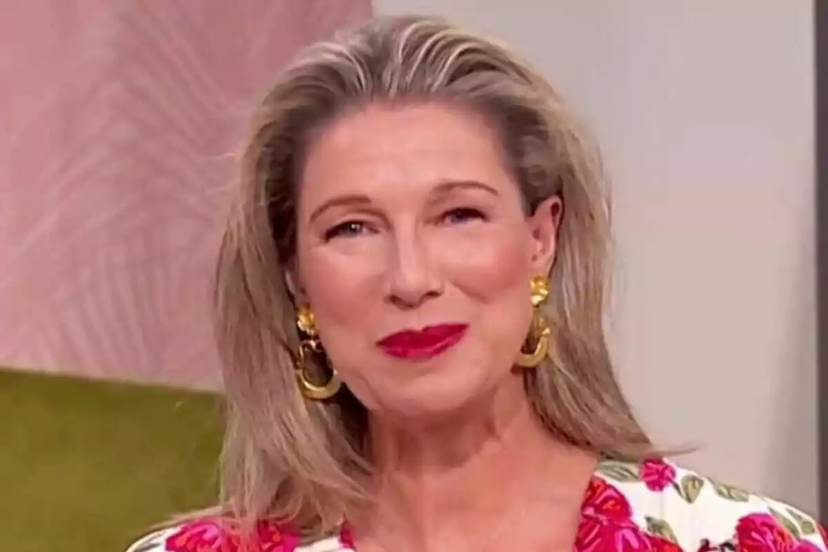 Anne Igartiburu de cabello rubio y largo con labios rojos, usando aretes dorados grandes y blusa con estampado floral, sonríe frente a un fondo claro en D Corazón
