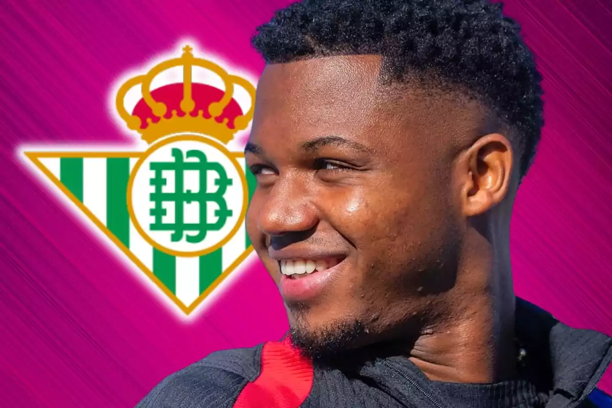 Un hombre sonriente con el escudo del Real Betis de fondo sobre un fondo rosa.