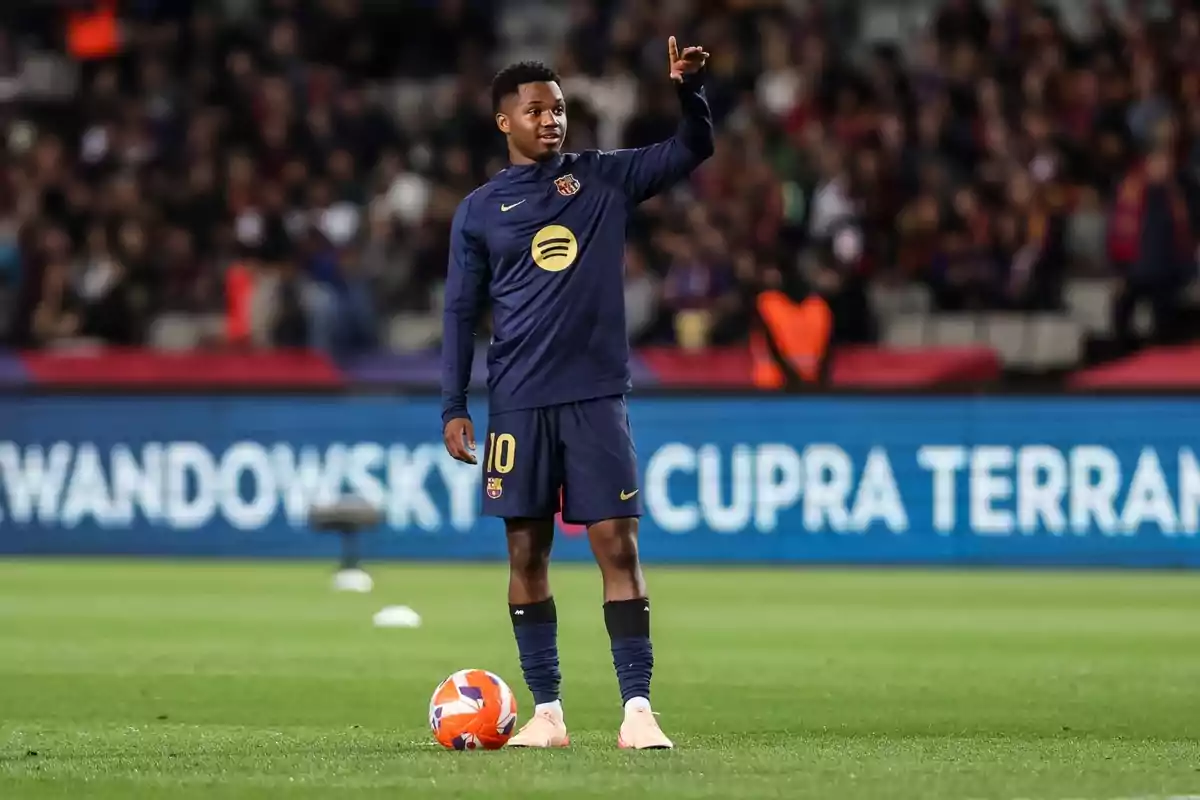 Jugador de fútbol con uniforme azul oscuro en el campo, levantando la mano y con un balón a sus pies. Jugador de fútbol con uniforme azul oscuro en el campo, levantando la mano y con un balón a sus pies.