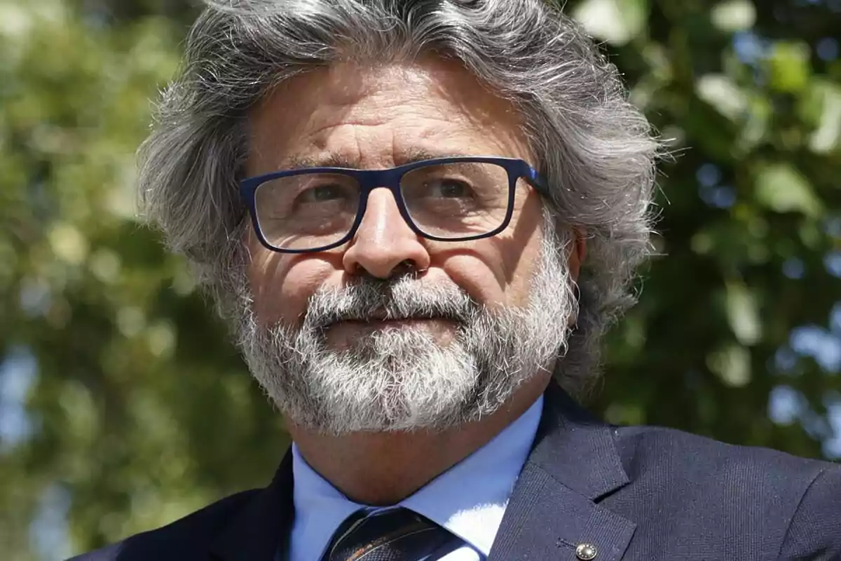 Hombre con barba y gafas azules, vestido con traje oscuro, al aire libre con fondo de árboles.