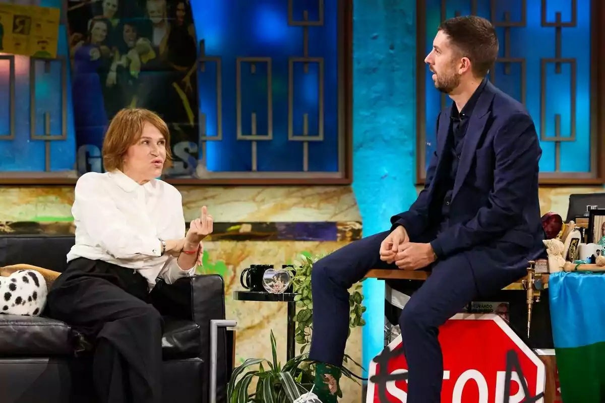 Antonia San Juan y David Broncano están sentados en La Revuelta, ella en un sofá negro y él en un taburete, ambos parecen estar conversando en un ambiente informal y colorido. Antonia San Juan y David Broncano están sentados en La Revuelta, ella en un sofá negro y él en un taburete, ambos parecen estar conversando en un ambiente informal y colorido.