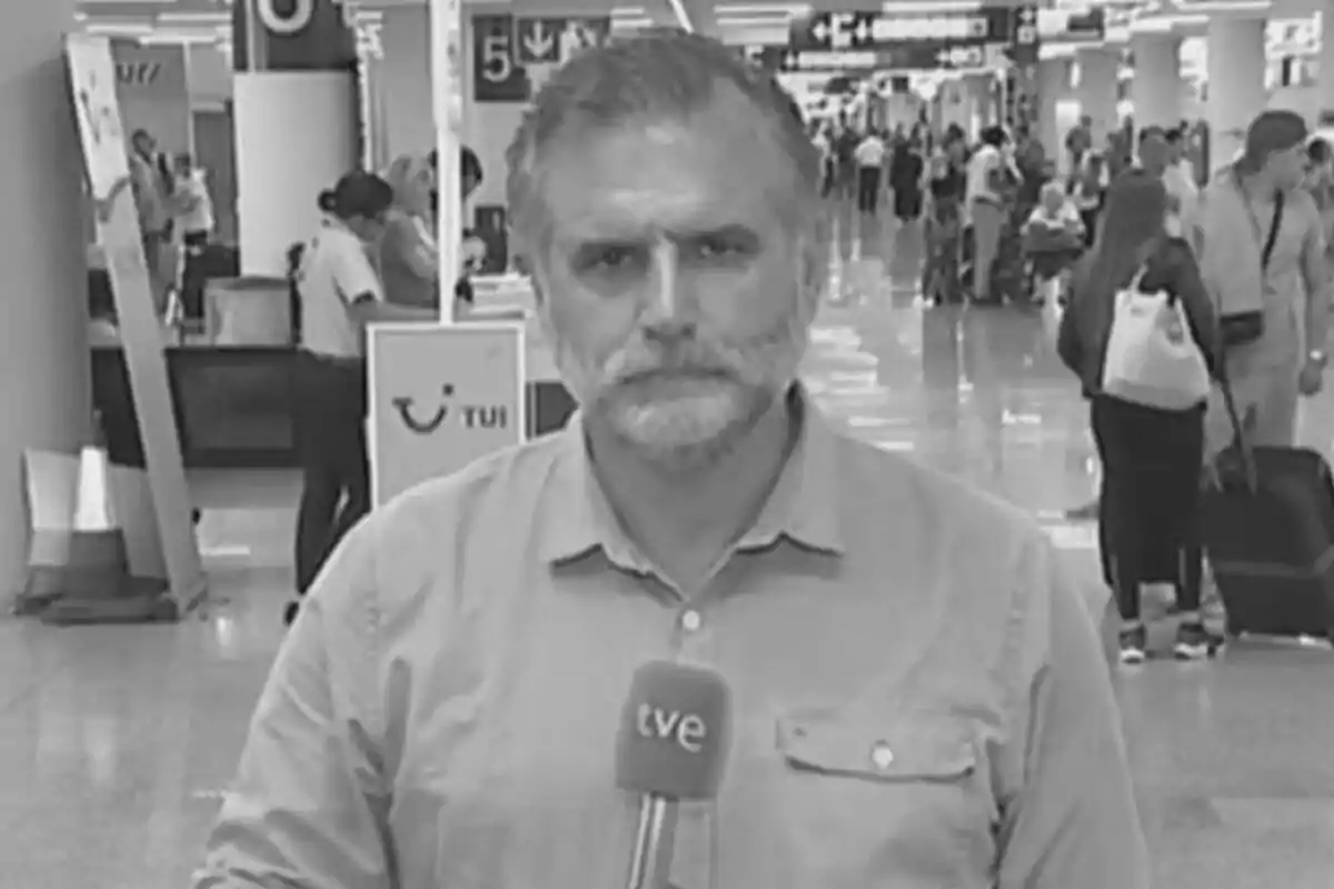 Fotografía en blanco y negro de Antonio Aranda con una camisa clara sostiene un micrófono de TVE en un aeropuerto concurrido.