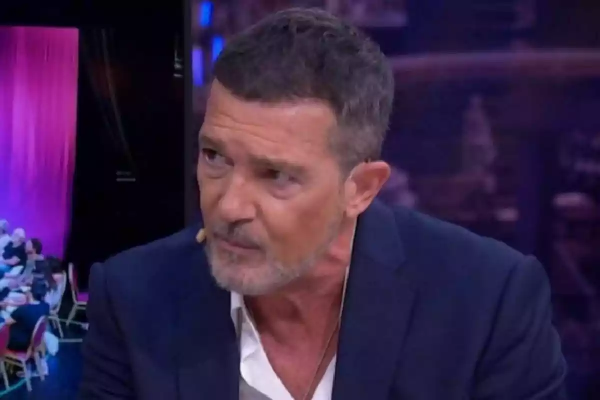 Antonio Banderas con barba y cabello corto, vestido con una chaqueta azul y camisa blanca, hablando en El Hormiguero con un micrófono en la oreja.