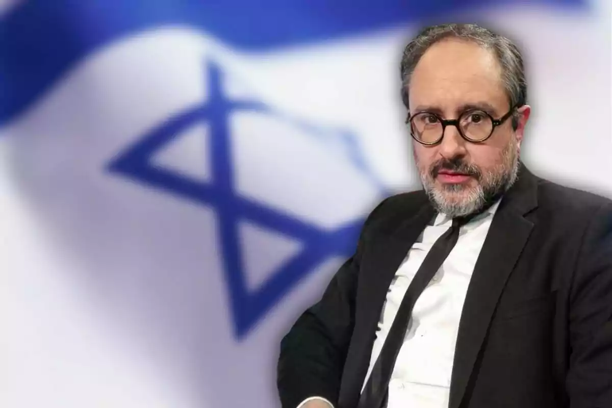 Hombre con gafas y traje oscuro frente a una bandera de Israel desenfocada.