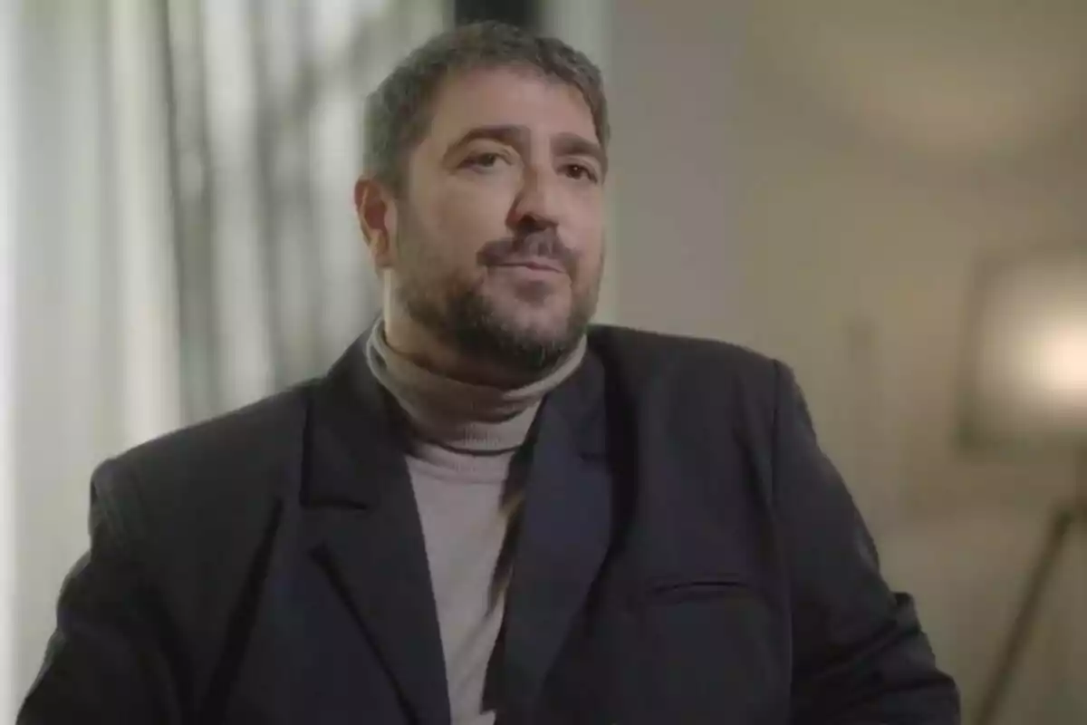 Antonio Orozco con cabello corto y barba, vistiendo un saco oscuro y suéter de cuello alto, sentado en un ambiente interior iluminado en 'Viajando con Chester'.
