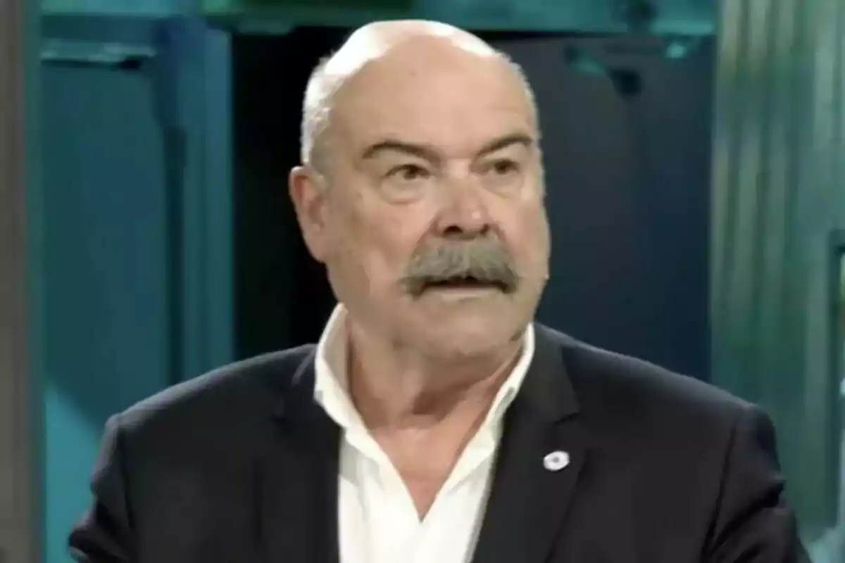 Antonio Resines de traje oscuro y camisa blanca con bigote y expresión seria en un entorno interior en La Revuelta. Antonio Resines de traje oscuro y camisa blanca con bigote y expresión seria en un entorno interior en La Revuelta.