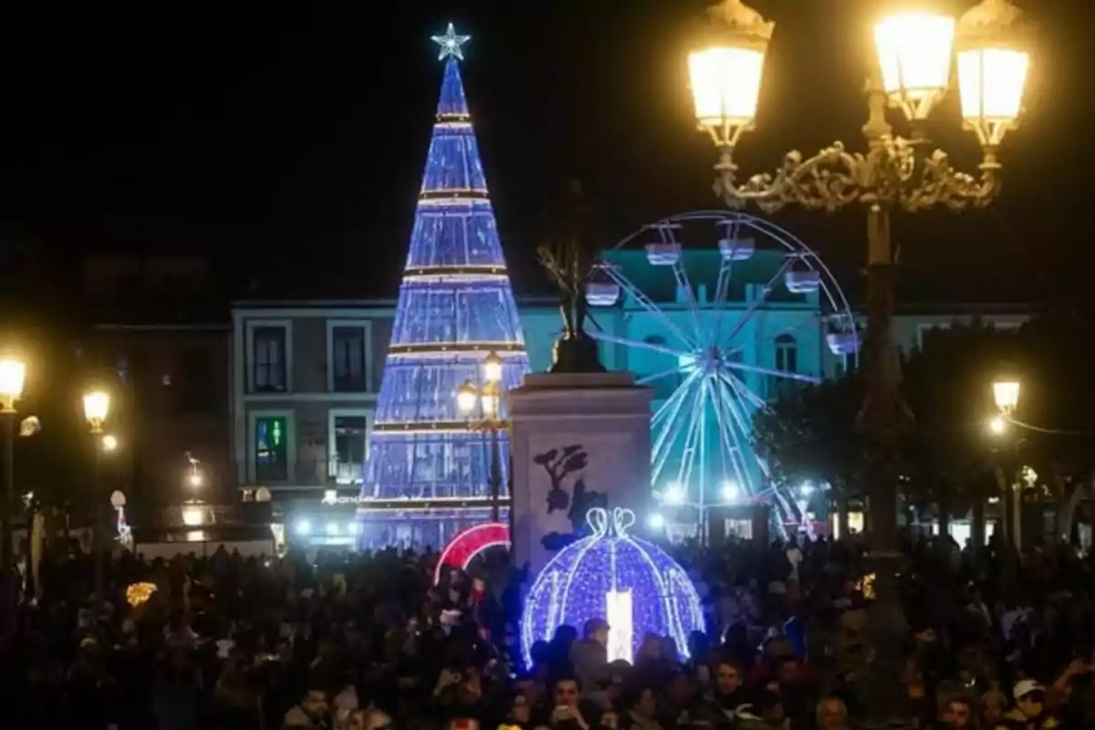 Una plaza iluminada por la noche con un árbol de Navidad azul, una noria y una multitud de personas disfrutando del ambiente festivo.