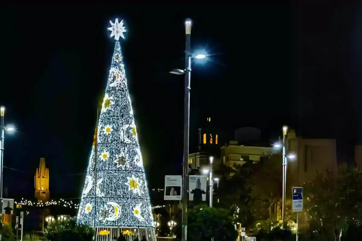 Un árbol de Navidad iluminado con decoraciones de estrellas y lunas en un entorno urbano nocturno.