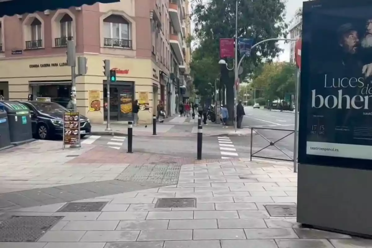 Una calle urbana con edificios, un semáforo en verde, un cartel publicitario y personas caminando por la acera.