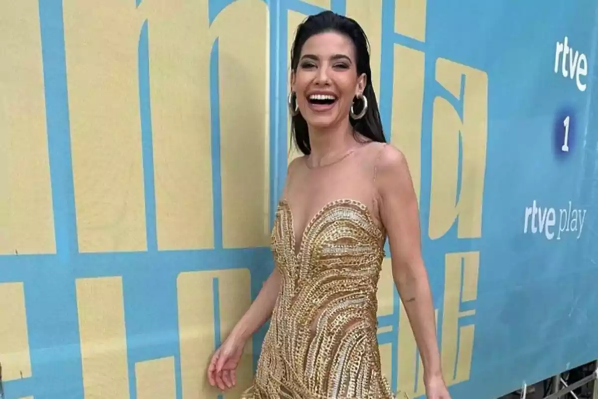 Ares Teixidó con un vestido dorado sonríe frente a un fondo azul con letras grandes en La Familia de la Tele. Ares Teixidó con un vestido dorado sonríe frente a un fondo azul con letras grandes en La Familia de la Tele.