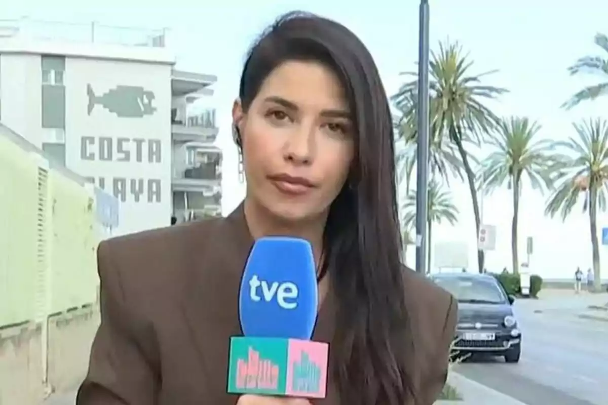 Ares Teixidó sostiene un micrófono azul de La Familia de la Tele con el logo de "tve" mientras está de pie en una calle con palmeras y un edificio al fondo que tiene un letrero que dice "Costa Playa".