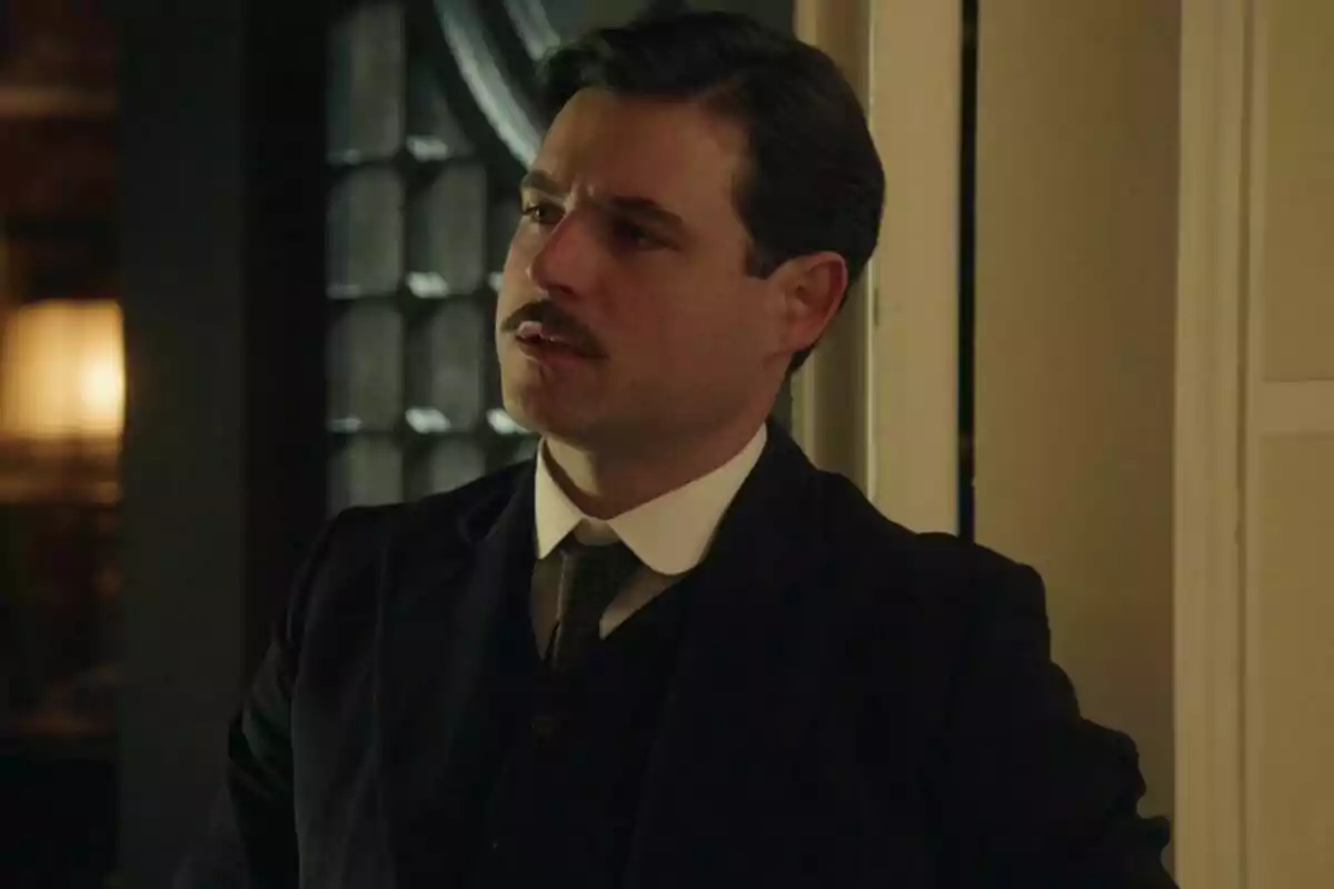 Arturo Sancho con bigote y traje oscuro está de pie junto a una puerta en un ambiente interior iluminado en La Promesa. Arturo Sancho con bigote y traje oscuro está de pie junto a una puerta en un ambiente interior iluminado en La Promesa.