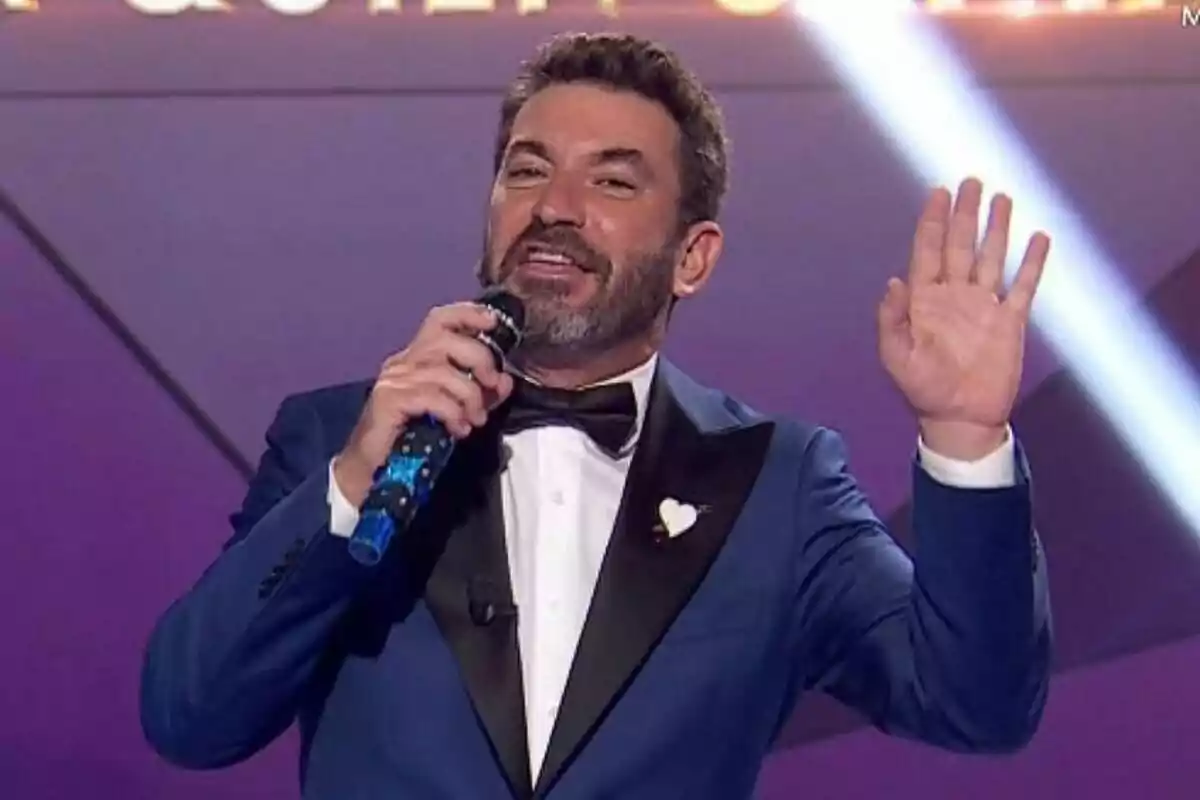 Arturo Valls con barba y traje azul sostiene un micrófono mientras levanta la mano en el plató de Mask Singer.