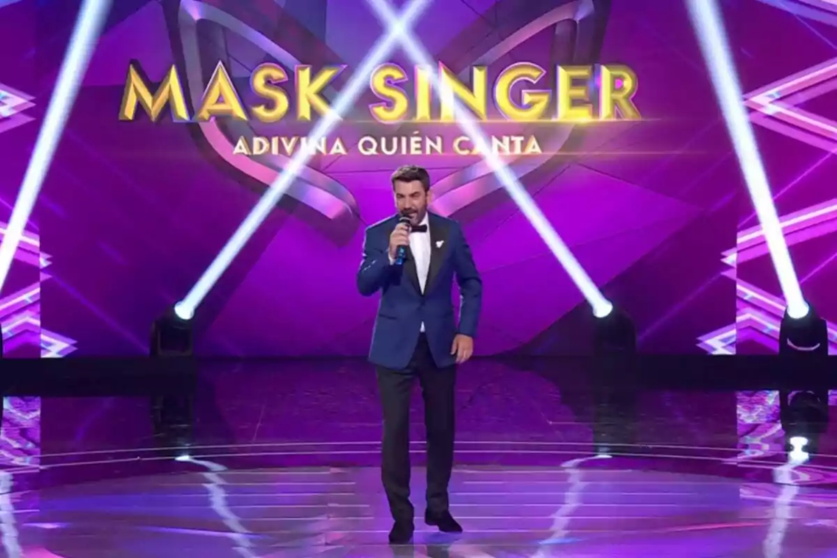 Un presentador con traje azul y pajarita negra sostiene un micrófono en el escenario del programa Un presentador con traje azul y pajarita negra sostiene un micrófono en el escenario del programa
