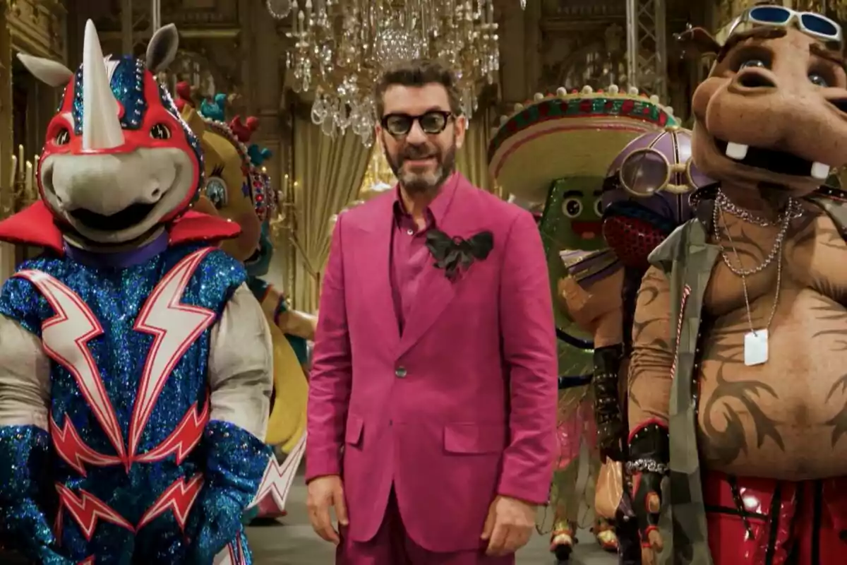 Arturo Valls con un traje rosa brillante está rodeado de personas disfrazadas de animales y personajes coloridos en un entorno elegante con candelabros en la promo de Mask Singer.