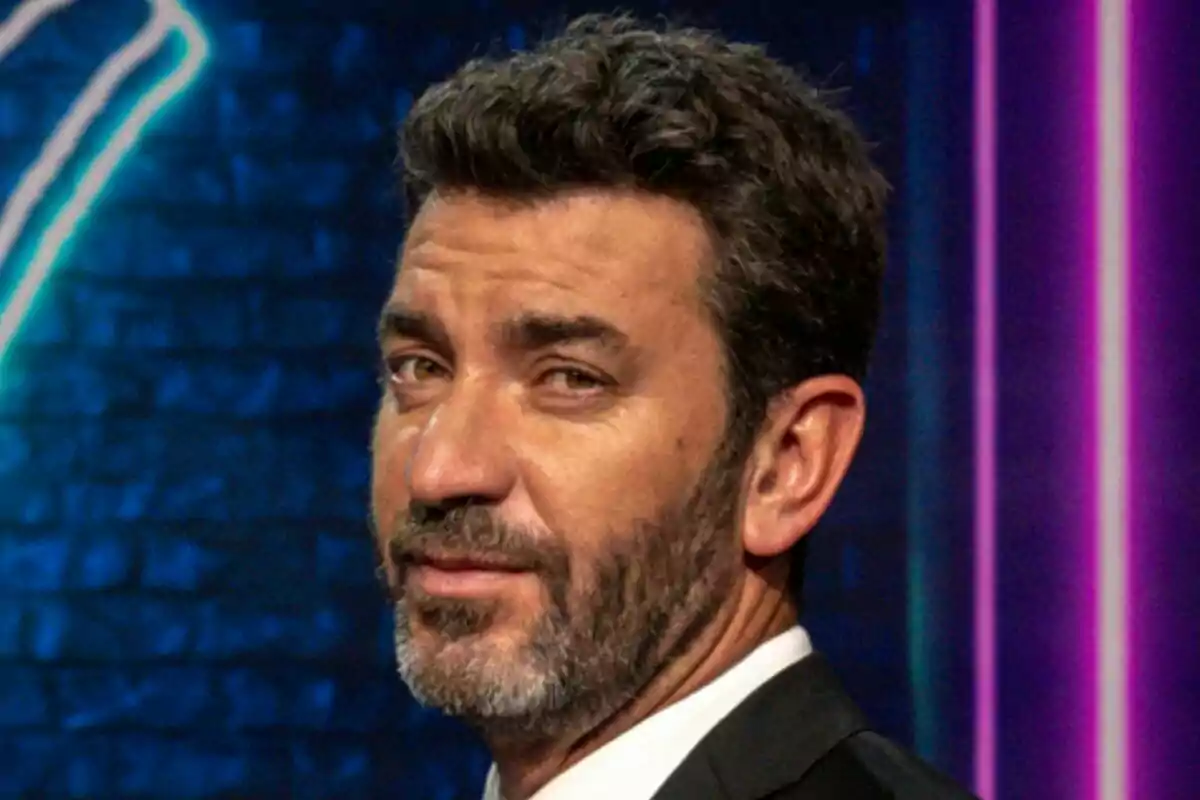 Arturo Valls con barba y cabello oscuro mira hacia la cámara con una expresión seria en un fondo oscuro con luces de neón en That's My Jam.