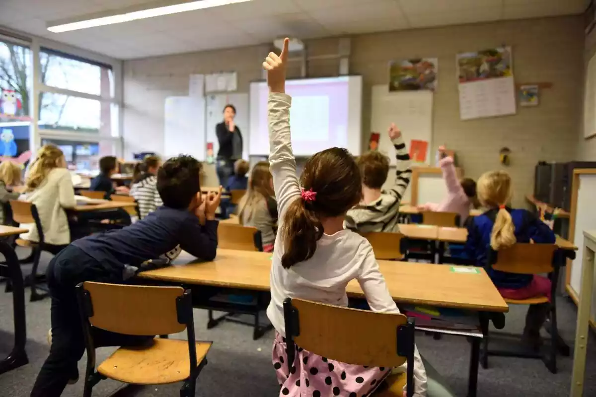 Un aula escolar vista por detrás con varios niños sentados en sus pupitres y una liña levantando su brazo izquierdo para preguntar a la profesora que hay en el fondo