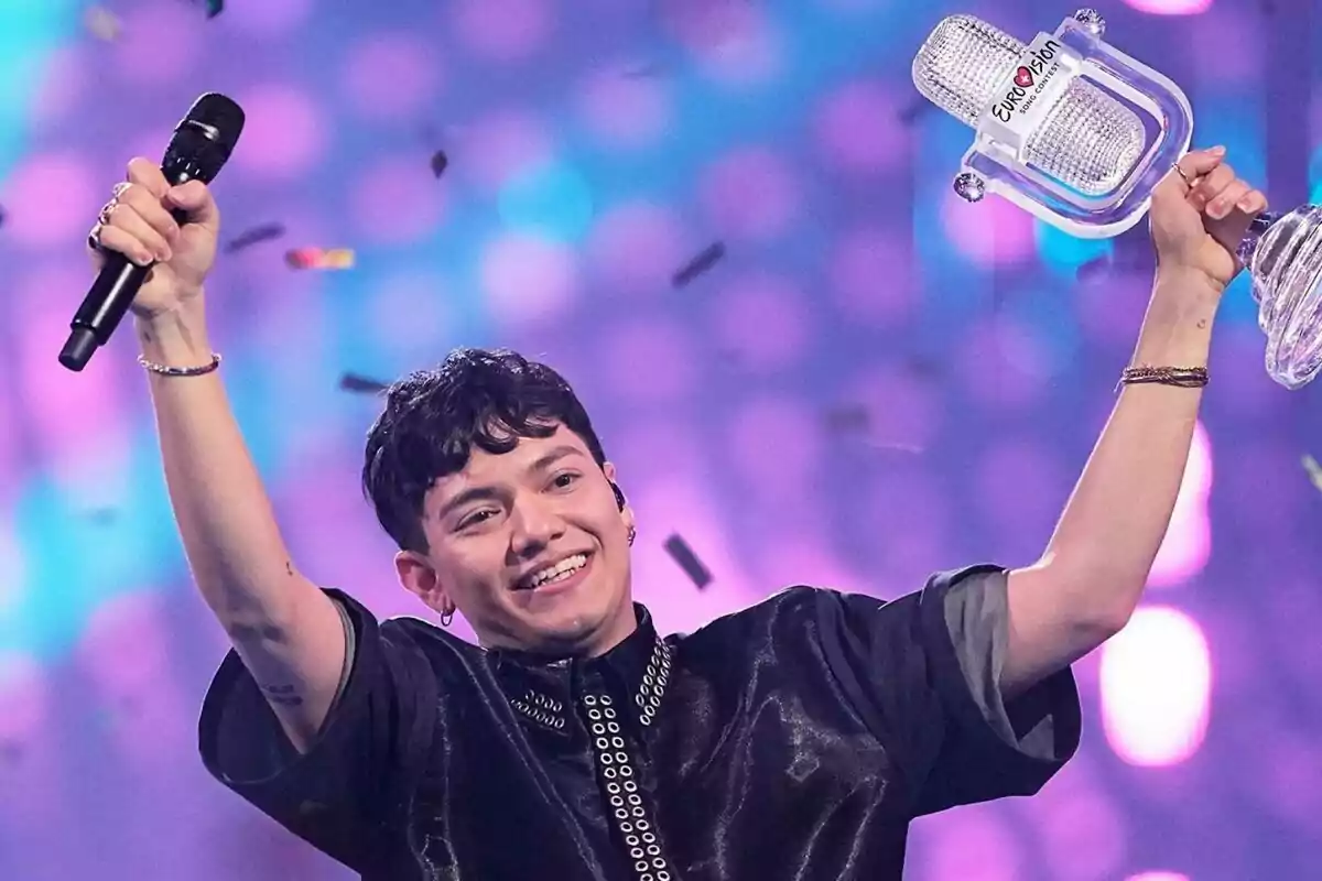 JJ sonriente sostiene un trofeo de micrófono de la victoria de Eurovisión por Austria y un micrófono en un escenario con luces de colores y confeti.