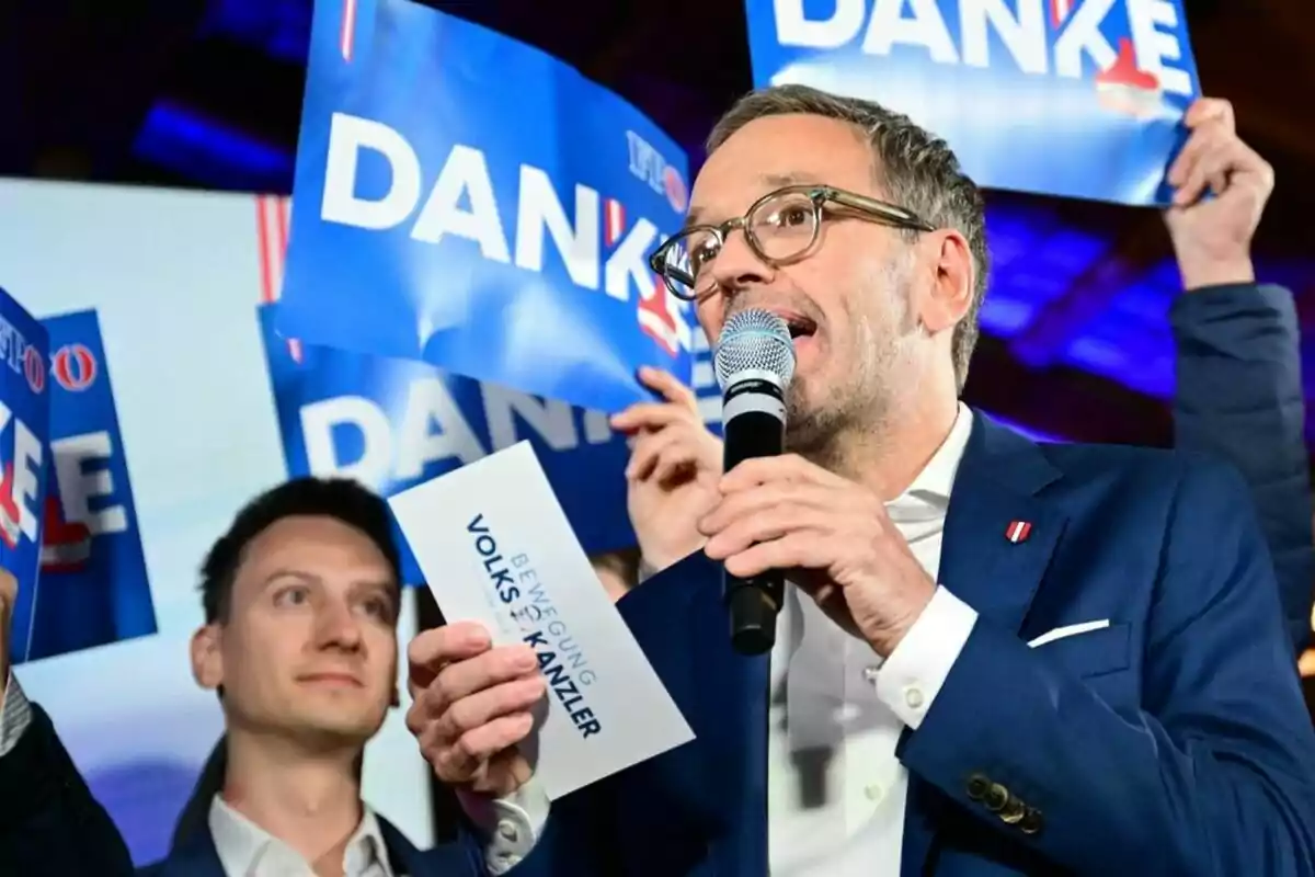 Un hombre con gafas y traje azul sostiene un micrófono mientras habla, rodeado de personas que levantan carteles azules con la palabra "DANKE".