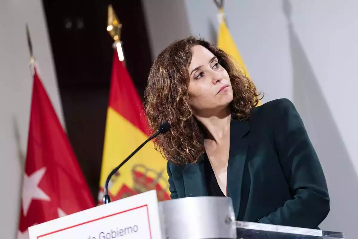 Fotografía de Isabel Díaz Ayuso de perfil, en un acto con las banderas de Madrid y España de fondo