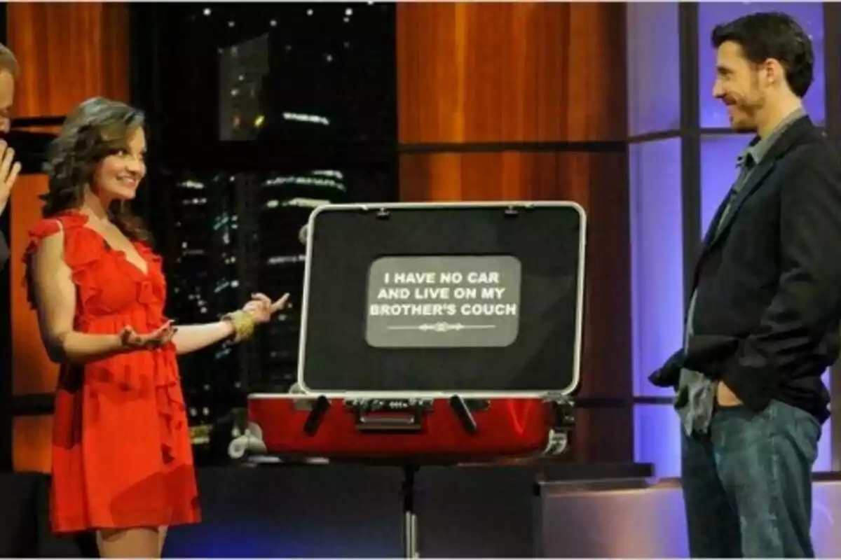 Una mujer con vestido rojo y un hombre con chaqueta se miran sonrientes en un set de televisión de Baggage junto a una maleta abierta que muestra un letrero en inglés que dice que no tiene auto y vive en el sofá de su hermano